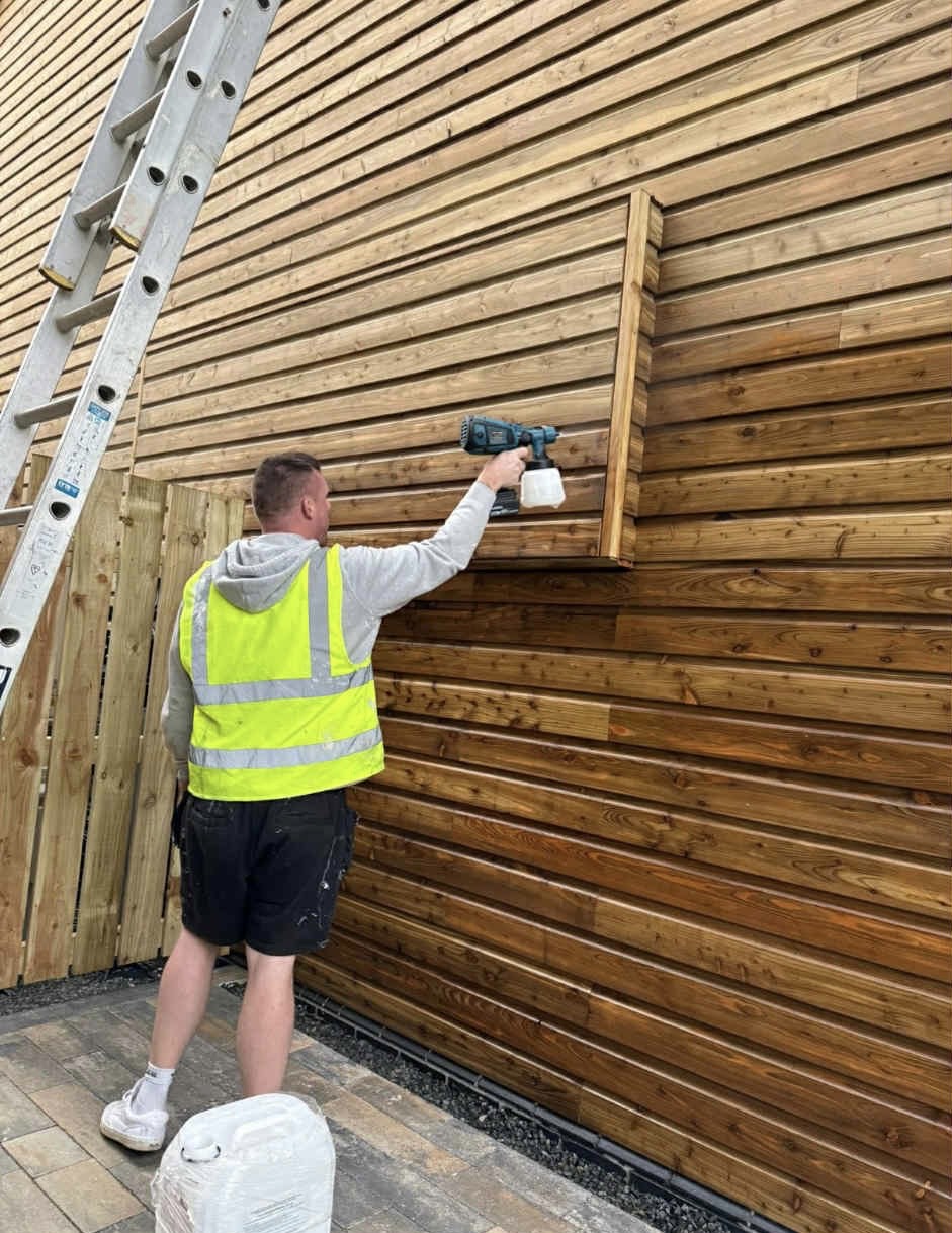 Painter spray painting wooden cladding on building exterior at Widemouth Bay